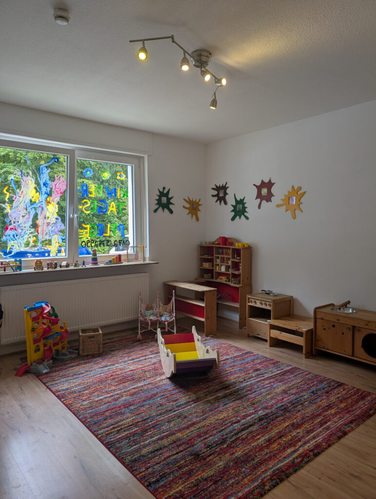 Helles Spielzimmer einer Kindertagespflege mit großem bunten Teppich, Holzspielzeug und kindgerechter Fensterdekoration