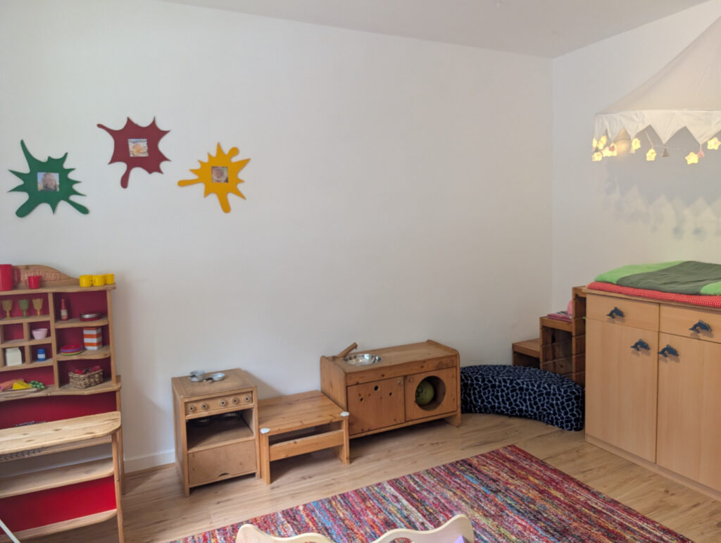 Helles Spielzimmer mit kindgerechten Holzmöbeln, einem roten Kaufladen, einer Spielküche und einem Wickeltisch mit Treppenaufgang.