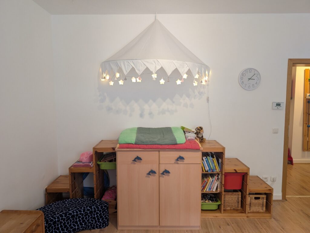 Gemütlicher Wickelbereich in der Kindertagespflege mit einer hölzernen Wickelkommode, einem Baldachin mit Lichterkette und umgebenden Bücherregalen.