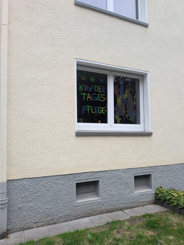 Fenster einer Kindertagespflege mit bunter, selbstgemalter Aufschrift 'Kindertagespflege' und Fensterbildern.