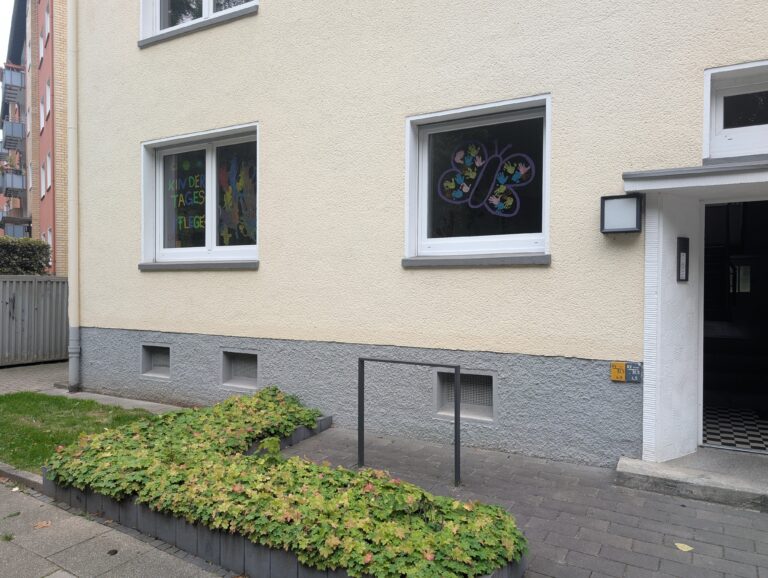 Beige Hausfassade einer Kindertagespflege mit bunter Fensterbeschriftung und einem Schmetterling aus Handabdrücken.