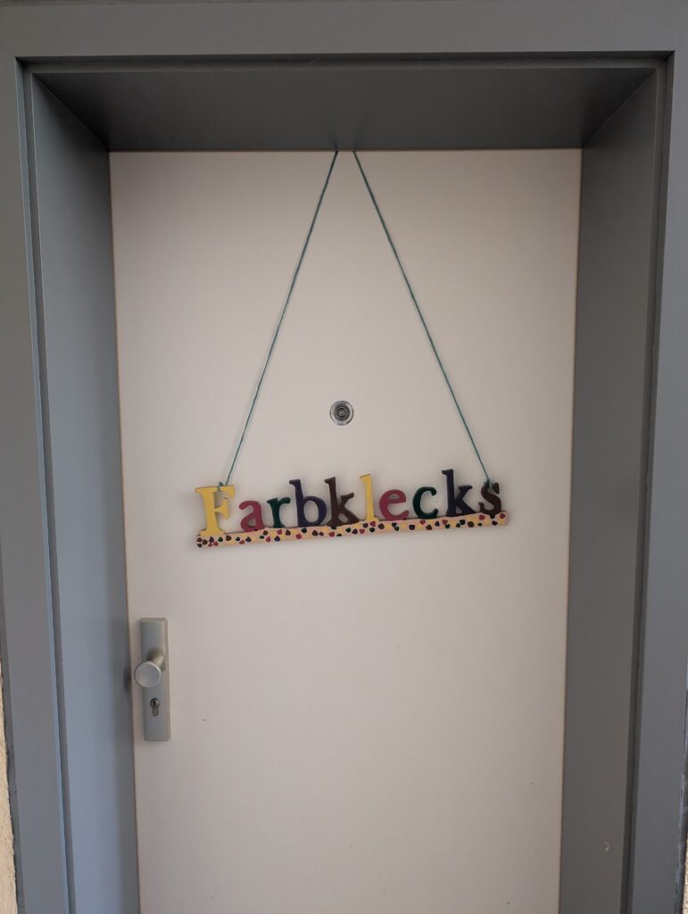 Weiße Zimmertür mit einem hängenden Holzschild mit der bunten Aufschrift Farbklecks.
