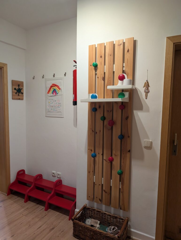 Kindgerechte Garderobe aus vertikalen Holzlatten mit bunten Haken, weißen Ablagen und roten Trittstufen im Flur.