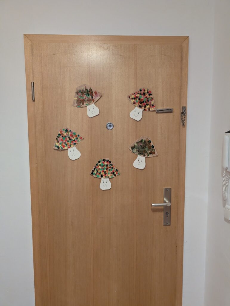 Helle Wohnungstür aus Holz, dekoriert mit fünf selbstgebastelten Papier-Pilzen mit bunten Punkten und lachenden Gesichtern.
