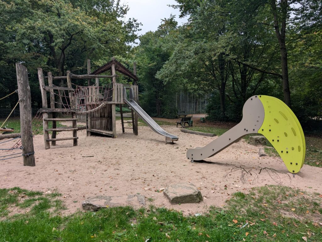 Spielplatz im Grünen mit Klettergerüst aus Holz und einer modernen Kleinkind-Rutsche im Sandkasten.