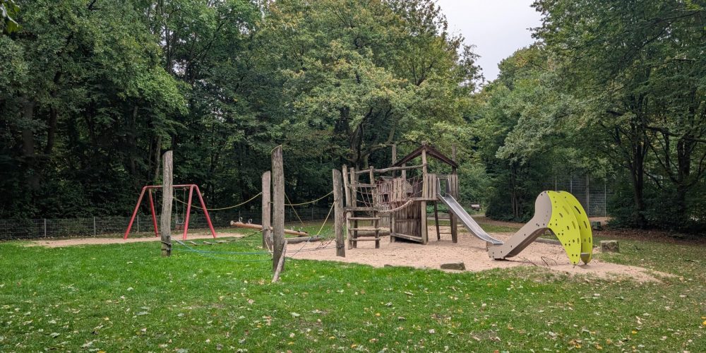 Leerer Spielplatz im Park mit hölzernem Klettergerüst, Rutsche und Schaukel auf einer Sandfläche umgeben von Bäumen.